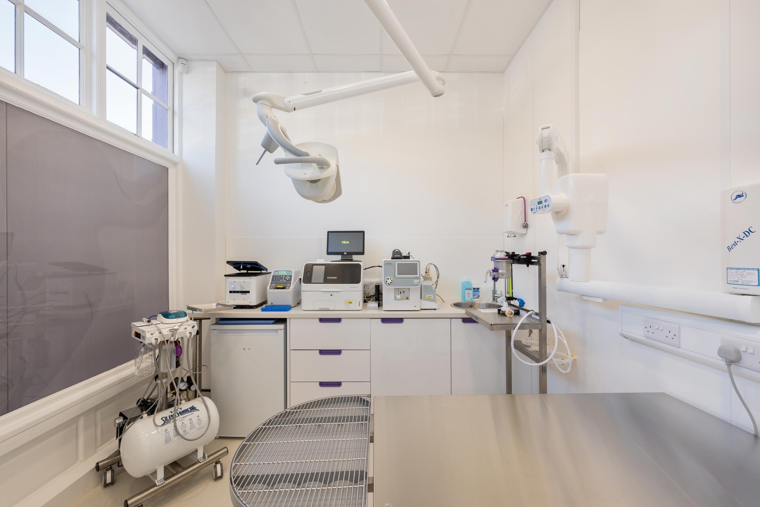 A modern veterinary clinic room featuring medical equipment, a treatment table, and bright daylight from large windows.