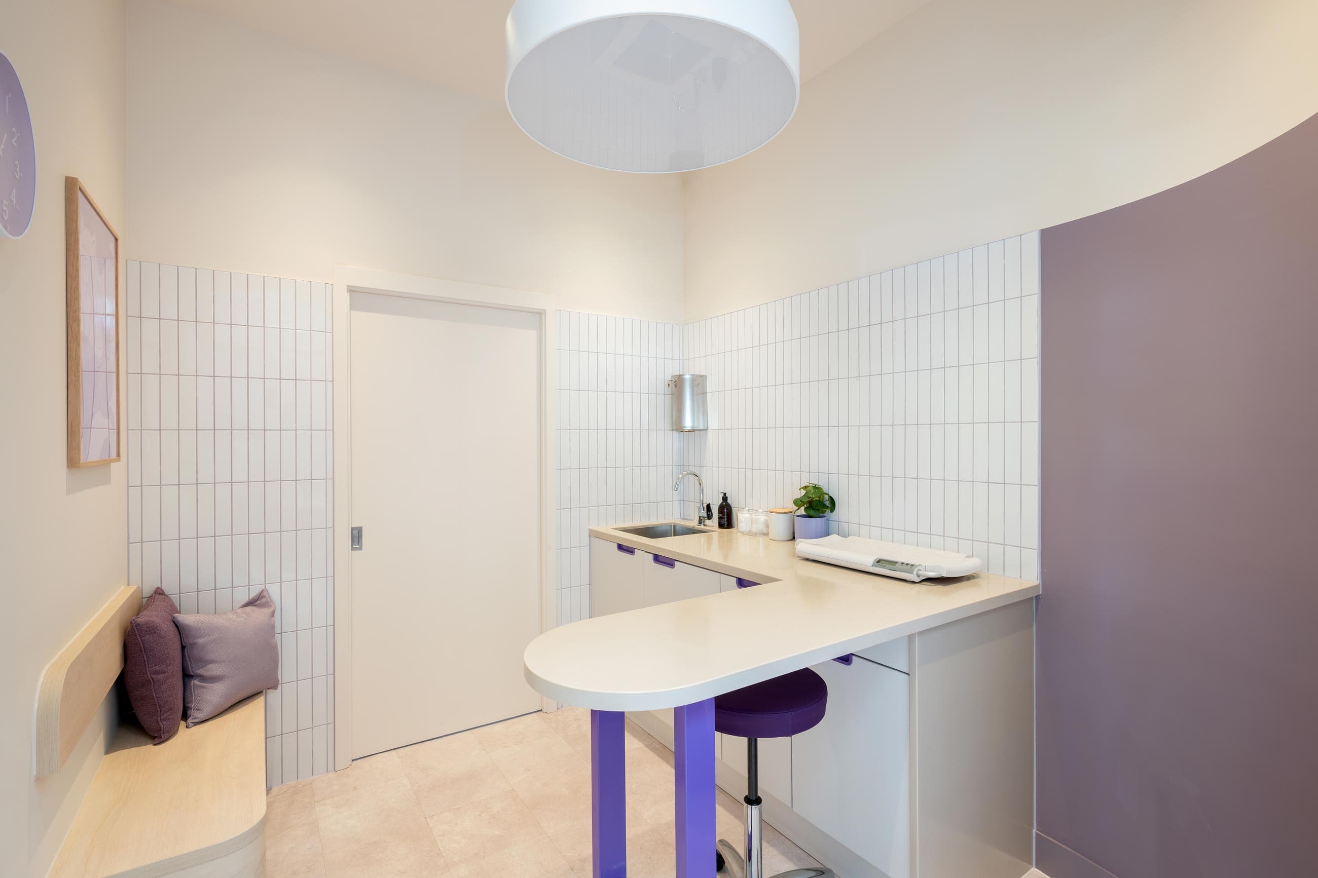 A modern, minimalistic clinic room featuring a white countertop, purple accents, tiled walls, and a comfy seating area.